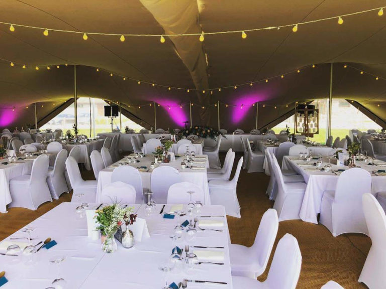 Des tables à manger rectangulaires sous un « tente stretch » orné de guirlandes lumineuses et d'un éclairage vers le haut de couleur violette