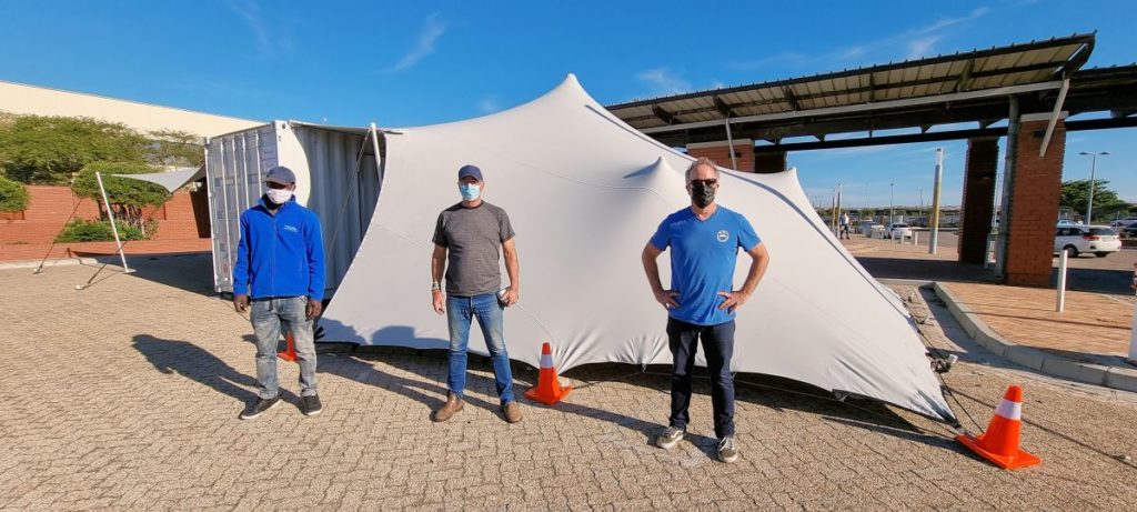 Het RHI-team naast een omgebouwde vaccinatiecontainer met een op maat gemaakte strektent als overkapping, waarvan één zijde is neergelaten