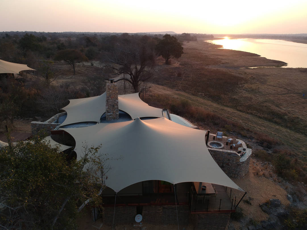Luftaufnahme des beigen „Luxury Custom Glamper Stretch“ in einem abgelegenen Camp am Wasser bei Sonnenuntergang in Sambia