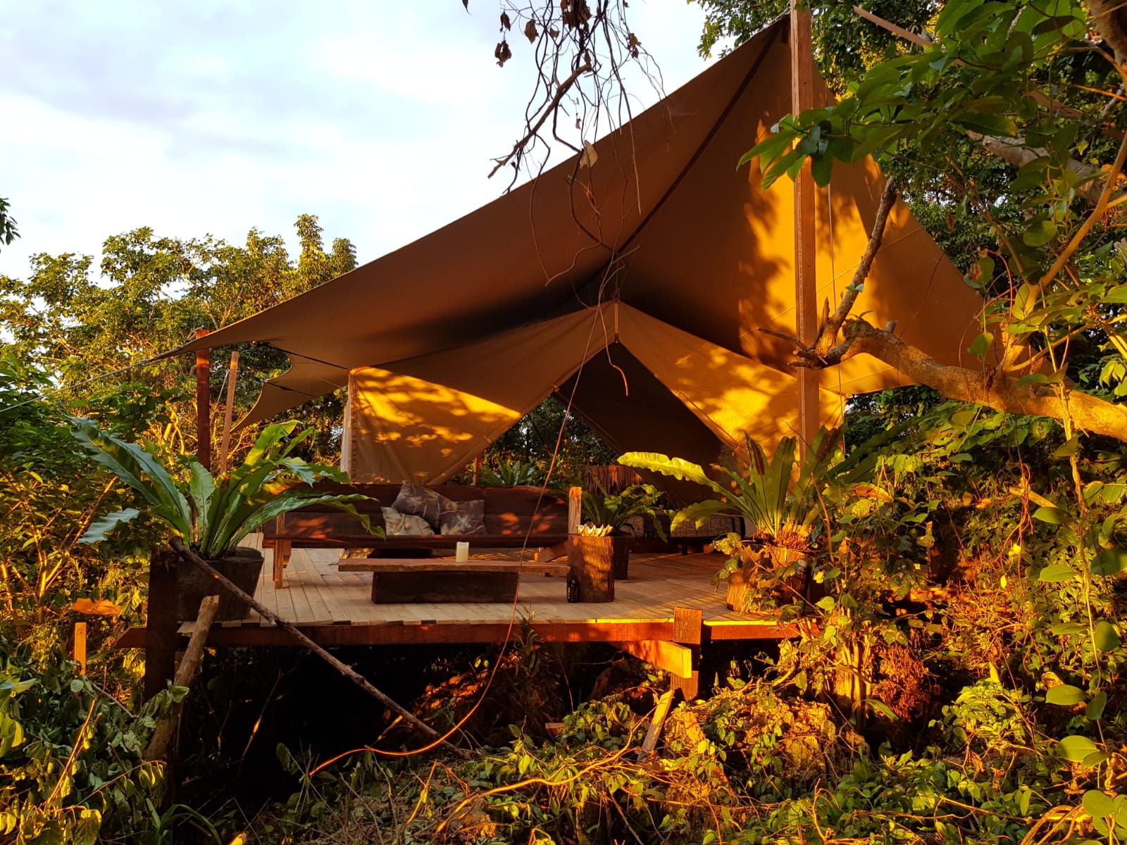 Beigefarbene „RHI Glamper“- Stretchzelt -Einheit, die eine Glamping-Terrasse aus Holz inmitten von Bäumen umgibt