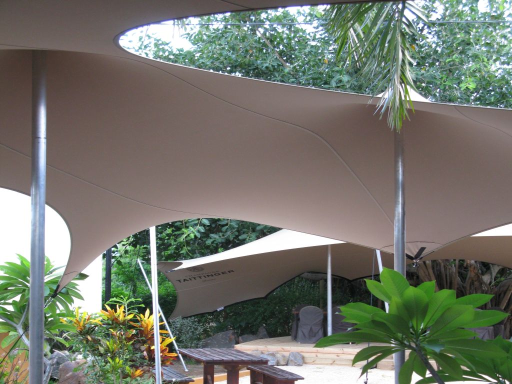 Geboogde, beige, grote Catenary-stretch-tent op maat boven een tuinterras met bomen en beplanting