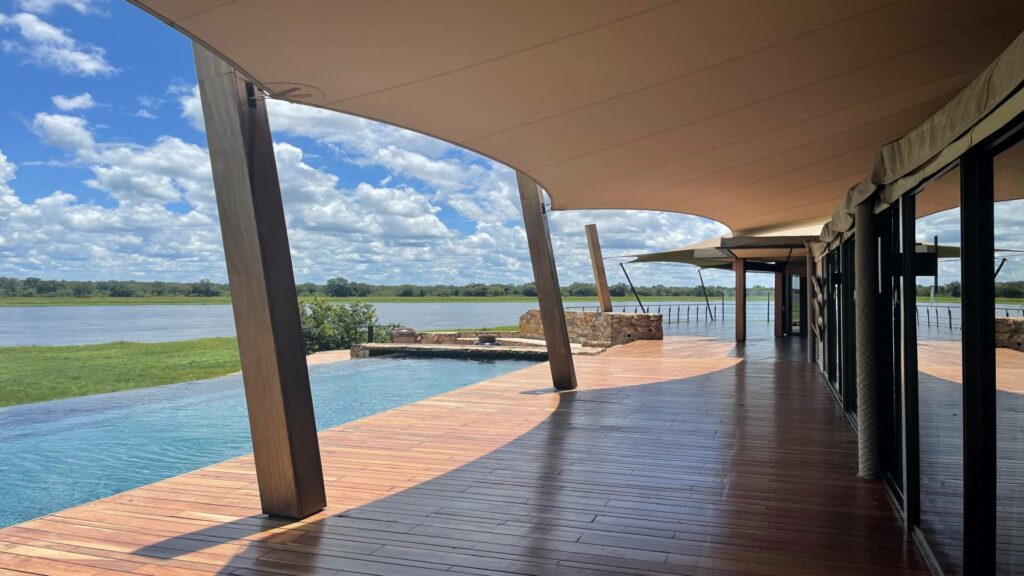 Beige, met kettingdraadrand afgewerkte, op maat gemaakte stretchtent boven een houten terras naast een zwembad en water, voor een luxe afgelegen kamp