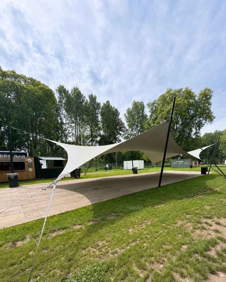 Light hypar stretch tent above a timber platform on a tree-lined lawn — 20 × 10 m Hypar Stretch Tent for a Festival in the Netherlands
