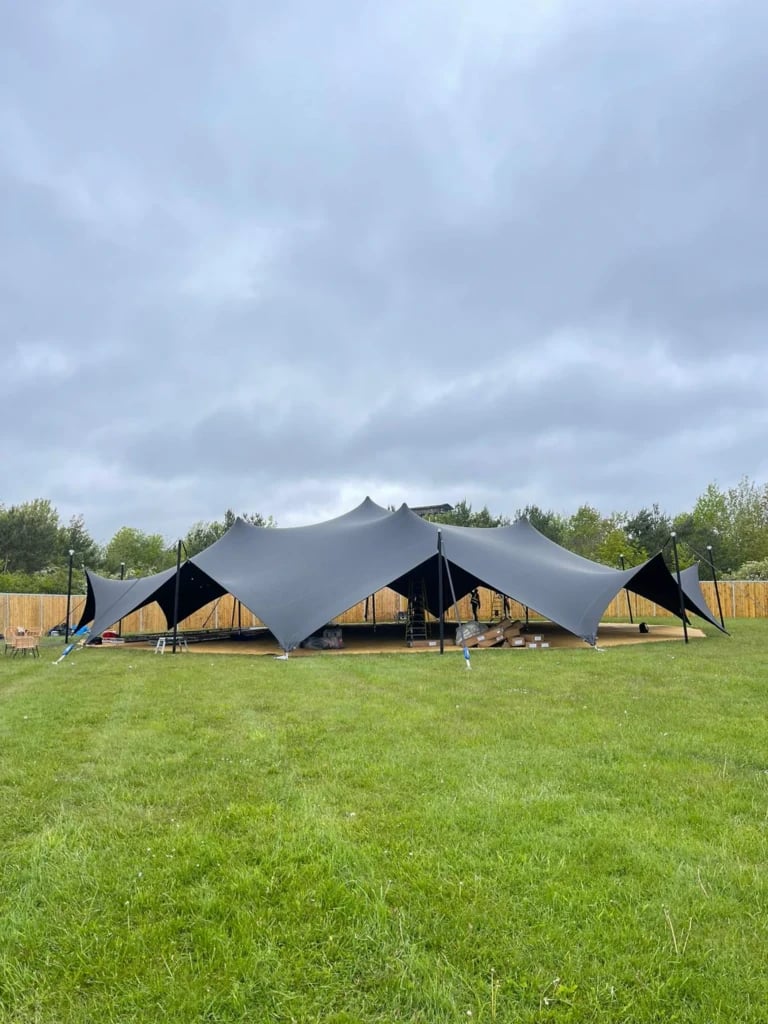 Ten-sided dark event tent above a timber platform on a lawn — Ten-Sided Arachnid Stretch Tent for BBC Festival in the UK