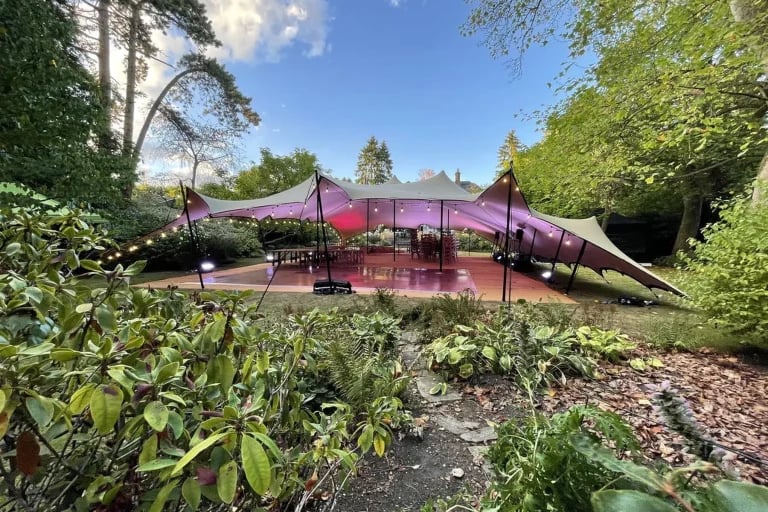 Vue d'ensemble, à travers les plantations du jardin, d'une installation « RHI » tente stretch mise en place à l'occasion d'un événement dans un jardin boisé